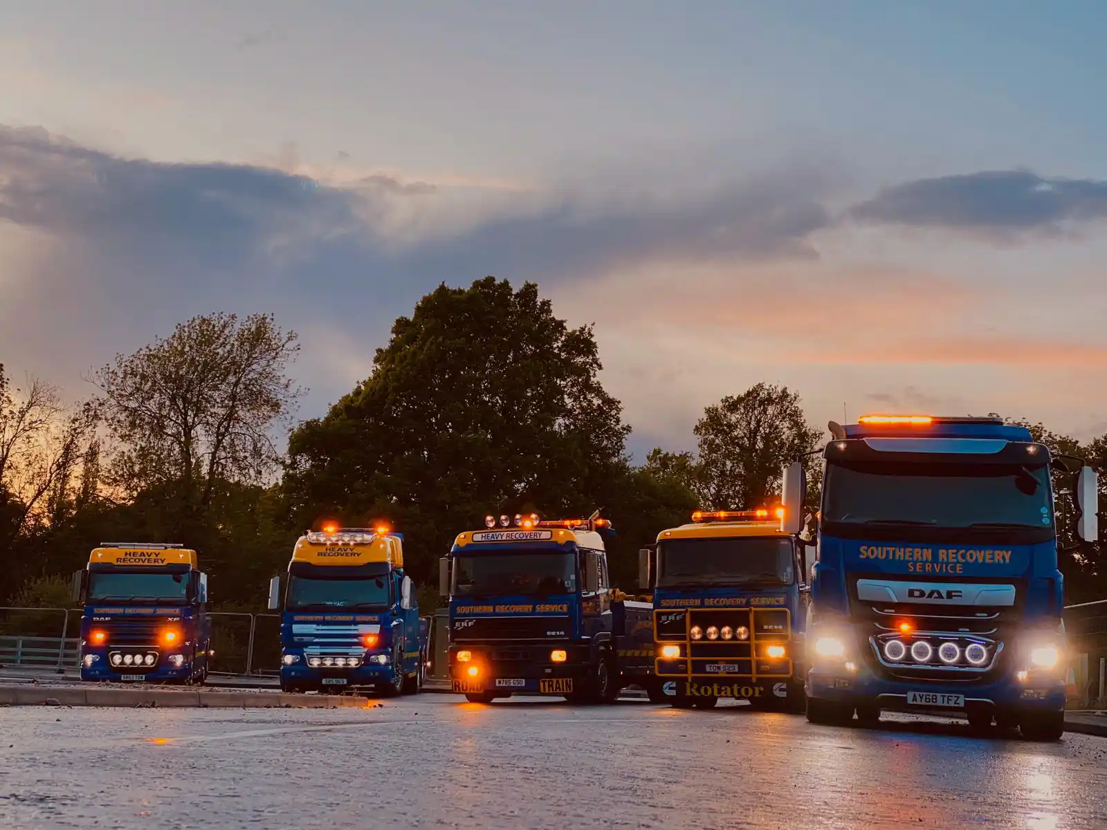 5 recovery trucks lined up in a row facing forwards with their lights on